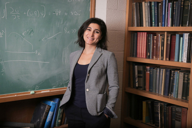 Lydia Bourouiba stands near a full bookshelf and chalk board.